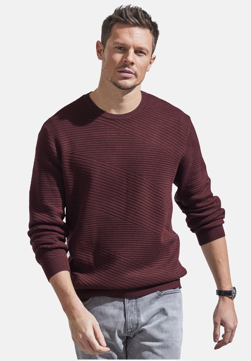 Burgunderfarbener Rundhals-Pullover mit diagonaler Rippstruktur, langen Ärmeln und gerippten Bündchen. Getragen über hellgrauen Hosen.