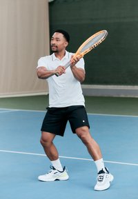 Jogador de ténis vestido com uma camisa polo branca e calções pretos, segurando uma raquete laranja, num campo azul com um fundo verde.
