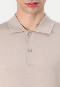 Polo en ribbed beige avec une texture douce, doté d'un col et de deux boutons transparents à la patte de boutonnage. Vue rapprochée.