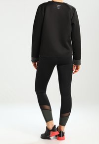 Czarna bluza z szarymi mankietami, z trzema poziomymi białymi liniami na plecach. Czarne legginsy z przezroczystymi panelami i oliwkowymi akcentami.