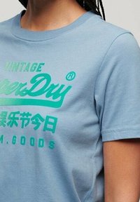 Ljusblå bomullströja med korta ärmar och rundad halsringning. Har djärv grön text och logotyp tryckt över framsidan i varierande storlekar.