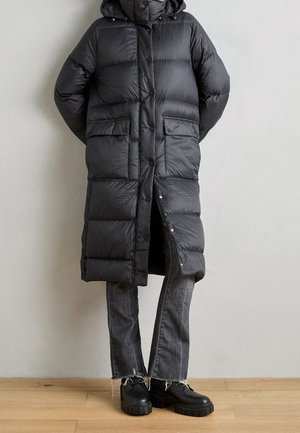 Personne portant un long manteau matelassé noir avec poches avant, un jean gris à ourlet effiloché et des chaussures noires à semelle épaisse, debout à l'intérieur.