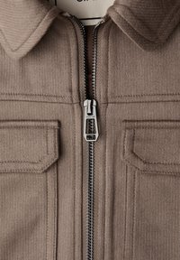 Brun manchesterjakke med spidst krave, sølv lynlås og to forlommer. Tekstureret stof og syningsdetaljer er synlige.