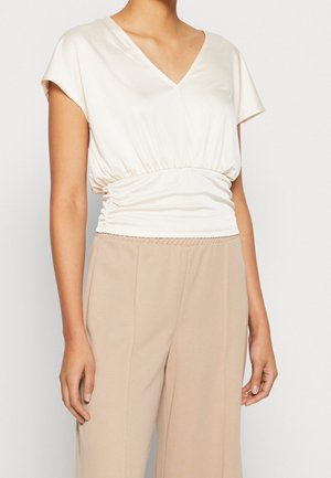 Haut beige clair avec décolleté en V, manches courtes drapées et détail de taille froncée, associé à un pantalon beige à large jambe en tissu doux.