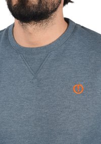Grå crewneck-tröja i mjukt material med en liten orange cirkellogo på vänster bröst samt ribbade muddar och halsringning.
