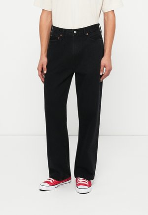 Black denim jeans with a relaxed fit, five pockets, and copper rivets, paired with red canvas sneakers. Visible seams and a plain design.