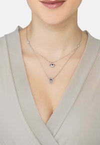 Silver layered necklaces featuring an eye charm and a hamsa hand, with blue accents and intricate detailing on each pendant.