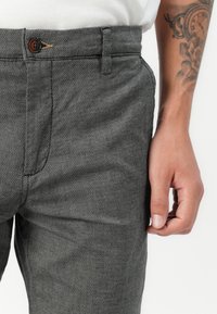 Grau gemusterte Hose mit normaler Taille, ausgestattet mit einem braunen Knopf und einem dezenten Webmuster. Hand auf Taillenhöhe platziert.