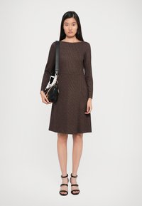 Braunes, langärmeliges Kleid mit tailliertem Oberteil und weit ausgestelltem Rock. Gewebte Textur. Kombiniert mit einer schwarzen Handtasche und schwarzen Riemchensandalen.