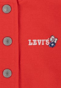 Rød bomuldsbluse med sølvknapper, "LEVI'S" logo i hvid broderi og blomsterdetaljer i blå og hvid. Blød tekstur.