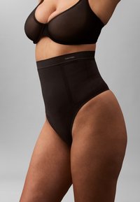 Kvinde iført sorte shaping-trusser med høj talje fra Calvin Klein og en matchet sort bh, vist fra midten af brystet til øvre lår.