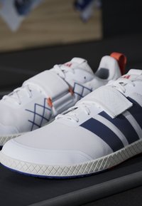 Biele športové topánky s modrými a červenými akcentmi, s textúrovaným povrchom, hrubou podrážkou a suchým zipsom cez hornú časť.