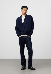 Marinblå stickad cardigan med sjalkrage, knappdetalj, bärs över en krämfärgad skjorta, matchad med mörk denimjeans och svarta skor.