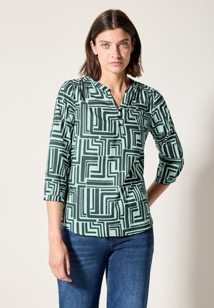 Blusa verde menta con motivo geometrico nero, scollatura a V, maniche a tre quarti e tessuto morbido. Modello indossata con jeans blu.
