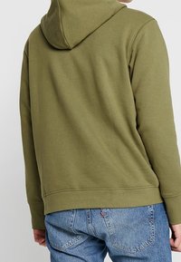 Persona con una felpa con cappuccio verde oliva e jeans di denim blu, vista da dietro, in piedi contro uno sfondo neutro.