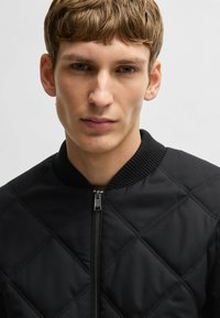 Black quilted bomber jacket with ribbed collar and zip closure, featuring a diamond-patterned texture and smooth fabric.