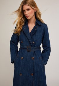 Dżinsowy trencz w ciemnoniebieskim kolorze, z paskiem w talii, podwójnym zapięciem, dużymi kieszeniami i brązowymi metalowymi guzikami.