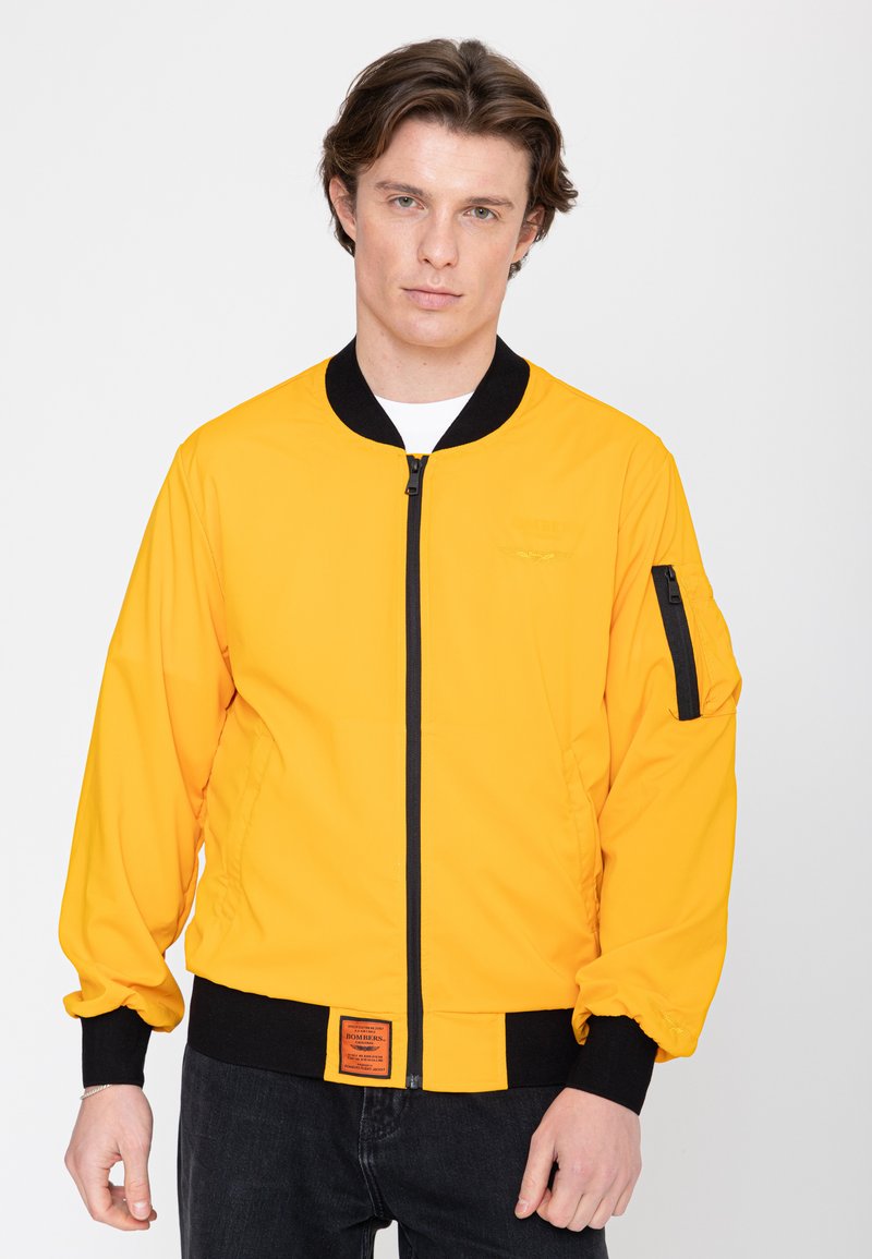 Junger Mann trägt eine leuchtend gelbe Zip-up-Jacke mit schwarzem Kragen, Bündchen und Saum, steht vor einem schlichten hellen Hintergrund.