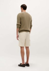 Shorts cargo beige clair avec poches latérales, associés à un pull en maille vert olive texturé. Manches visibles et chaussures brunes décontractées.