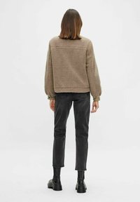 Persona in piedi di spalle, indossando una giacca in pile beige, jeans neri a vita alta e stivaletti neri su uno sfondo bianco.
