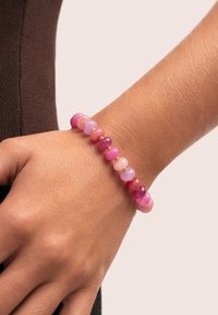 Pulsera de cuentas con piedras redondas de color rosa, púrpura y rojo con acentos dorados, llevada en una mano sobre un fondo de tela marrón.