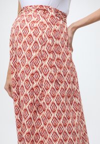 Jupe maxi en tissu léger, présentant un motif géométrique rouge rosé et crème, avec une taille nouée et un détail de poche latérale.