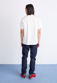 Witte katoenen t-shirt met korte mouwen, gecombineerd met donkerblauwe spijkerbroek. Rode sportschoenen met zwarte accenten. Achteraanzicht.