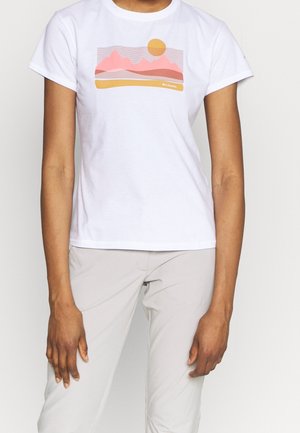 Person trägt weißes kurzärmeliges T-Shirt mit pinker und orangener Berg- und Sonne-Grafik, kombiniert mit hellbeiger Hose.