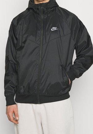 Man die een zwarte Nike vest met ritssluiting en capuchon draagt met zakken en een witte broek, staand tegen een effen achtergrond.