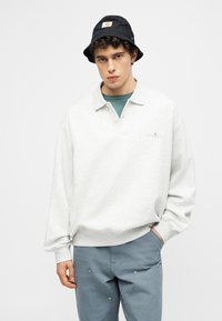 Νέος άνδρας που φοράει μαύρο καπέλο τύπου bucket, λευκό πλεκτό πόλο με μακριά μανίκια, τιρκουάζ εσωτερικό πουκάμισο και μπλε παντελόνι, με το ένα χέρι στην τσέπη.