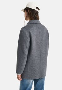 Grijze wollen jas met een gestructureerd ontwerp, geknipt kraag en rechte snit. Bevat een rugnaad en wordt gecombineerd met een witte pet en blauwe spijkerbroek.
