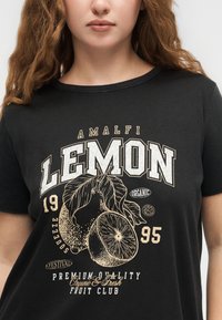 Κοντινό πλάνο ατόμου που φοράει μαύρο μπλουζάκι με γραφικό vintage λεμονιού και κείμενο "Amalfi Lemon 1995 Premium Quality Organic & Fresh."