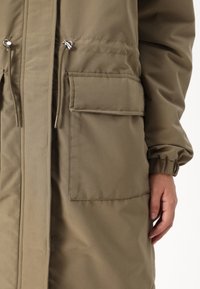 Khaki parka se zipem vpředu, stahovacími šňůrkami v pase, elastickými manžetami a dvěma velkými kapsami vpředu. Hladká, odolná tkanina.