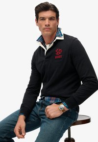 Zwart rugbyshirt met lange mouwen en witte kraag, met daaroverheen een blauw-groene geruite blouse, gecombineerd met denim jeans.