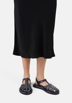 Voeten met zwarte leren sandalen met studs, gecombineerd met een zwarte midi-rok, afgebeeld tegen een effen witte achtergrond.
