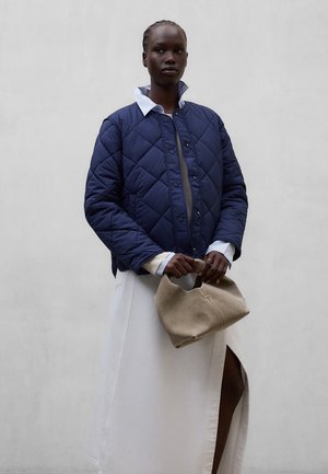 Mujer con chaqueta acolchada azul marino sobre una camisa blanca con cuello y una falda blanca larga con una abertura, sosteniendo un bolso blando beige.