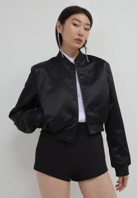 Frau trägt lange Strass-Ohrringe, eine schwarze Satin-Bomberjacke, ein weißes Button-Up-Hemd und schwarze hochgeschnittene Shorts vor einem weißen Hintergrund.