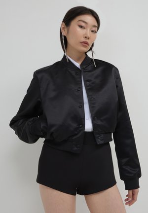 Frau trägt lange Strass-Ohrringe, eine schwarze Satin-Bomberjacke, ein weißes Button-Up-Hemd und schwarze hochgeschnittene Shorts vor einem weißen Hintergrund.