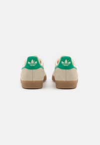 Beige suede sneakers with green accents and a textured gum sole, featuring a classic three-stripe logo on the heel.