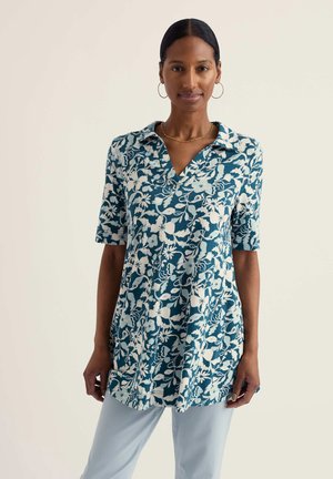 Naine, kes kannab sinise ja valge lillemustriga lühikeste varrukatega kraega tuunikat ja helesiniseid pükse, seisab ühevärvilise beeži tausta ees.