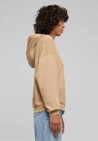 Sudadera beis con un bolsillo frontal, ajuste holgado, puños acanalados y dobladillo redondeado, combinada con jeans de lavado claro. Textura suave.