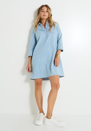 Robe bleu clair avec broderies florales, col montant et manches trois-quarts, accompagnée de baskets blanches.
