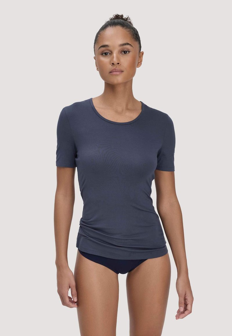 Femme avec les cheveux en chignon, portant une chemise ajustée à manches courtes bleu marine et des sous-vêtements assortis, debout devant un fond clair uni.
