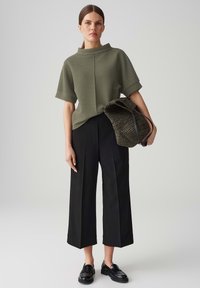 Top olivastro a maniche corte con colletto alto, abbinato a pantaloni neri a gamba larga. La modella tiene in mano una borsa verde scuro texture con manico in pelle.