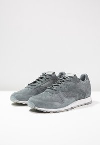 Une paire de baskets en suede gris de Reebok avec des lacets, une zone de orteil perforée et des semelles en caoutchouc blanches sur une surface blanche.