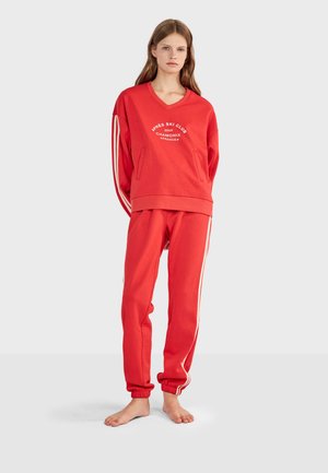 Jeune femme debout pieds nus portant un sweat rouge avec un texte blanc et un pantalon de jogging rouge assorti avec des bandes blanches sur les côtés.