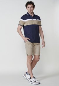 Tengerészkék és bézs csíkos pólóing, gallérral, khaki sorttal párosítva. A modell fehér és szürke sportcipőt visel kék akcentussal.