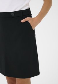 Jupe noire avec une fermeture à boutons sur le devant, des poches latérales et une texture lisse. L'ourlet est droit et tombe au-dessus du genou.