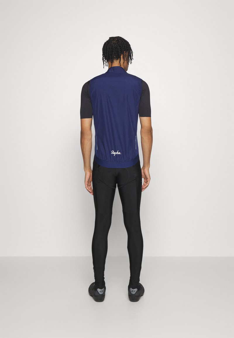 Marineblauwe fietsvest met hoge kraag, lichtgewicht materiaal, subtiele branding aan de zoom, in combinatie met zwarte, aansluitende leggings en schoenen.