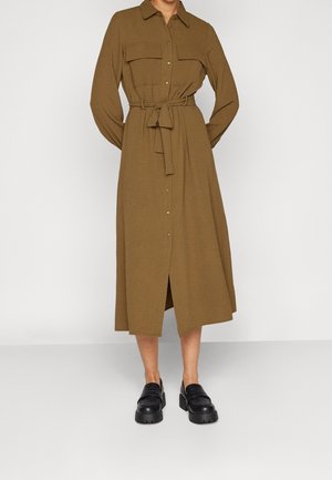 Femme portant une robe midi marron à manches longues, boutonnée sur le devant avec une ceinture à la taille et des mocassins noirs épais, debout devant un fond uni.